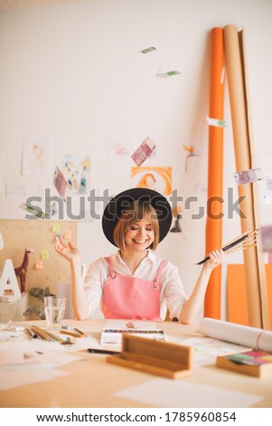 Creative smiling girl illustrator sitting under rain of money in workshop. Concept photo about art and freelance