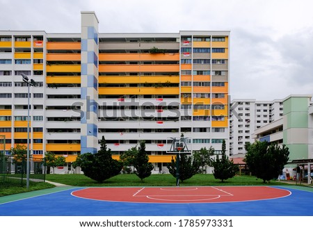 Colourful block of HDB flats (painted in white yellow orange blue) in yishun on bright, clear day, Singapore flags hung over some balconies. Neighbourhood park and basketball court in front. Royalty-Free Stock Photo #1785973331