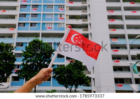 Hand waving Singapore flag outside on sunny day, against background of HDB flats and trees foliage. Many residents hang Singapore flags over balconies. Yishun Royalty-Free Stock Photo #1785973337