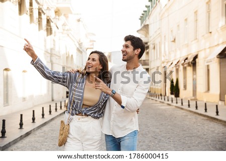 Image of young caucasian brunette couple smiling and pointing fingers aside while walking on city street