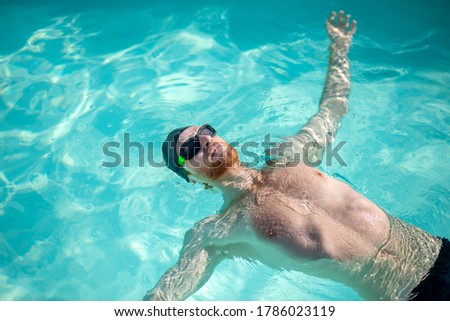 Lying on water. Bearded swimmer in black swimming cap and dark swimming goggles lying in water on his back