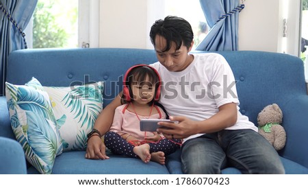 The young Asian father sat on the couch with his daughter while watching a movie with a smartphone                          