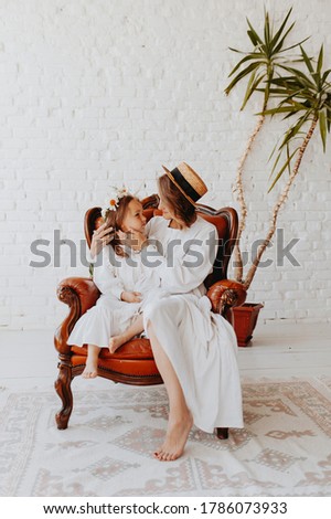 mom and daughter in a white room sitting on a brown chair dressed in white dresses, emotional and funny family photos, mom dressed in a straw hat