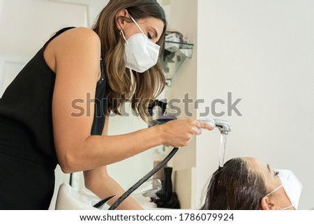 Beautiful and elegant young hairdresser rinsing with water the head wash of a young client with long brown hair, in the hairdresser's shop. Social distancing. Use of the face mask. Close-up Royalty-Free Stock Photo #1786079924