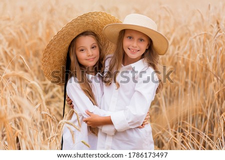 Cute little girls sisters with blond hair in a summer field at sunset in white dresses with a straw hat. Joyful girlfriends enjoy summer in the field
