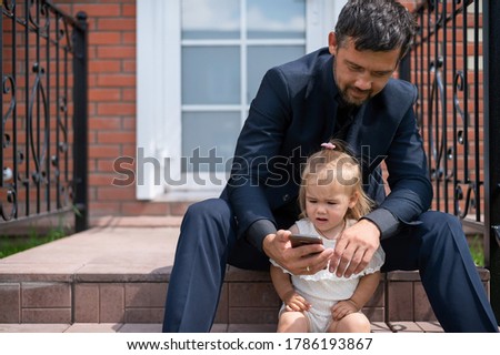 Caucasian man in a business suit looks at the mobile screen with daughter on the stairs. A little girl with a loving father watching cartoons on a smartphone while sitting on the porch of the house.