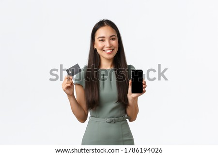 Small business owners, women entrepreneurs concept. Smiling good-looking asian woman in dress showing mobile screen application and credit card, introduce banking app, white background