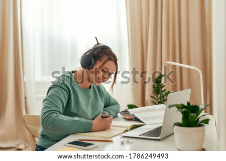 Studying online. Young beautiful woman, happy female student wearing headphones, listening online course and making notes while sitting at desk. Online study. Distance education. E-learning