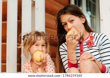 Two little girls in the summer at the dacha eat green apples while sitting on a bench. Happiness and fun concept. Summer holidays.