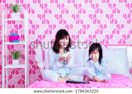 Portrait of mother and her daughter looks sick while wiping their nose with tissue and sitting on the bed