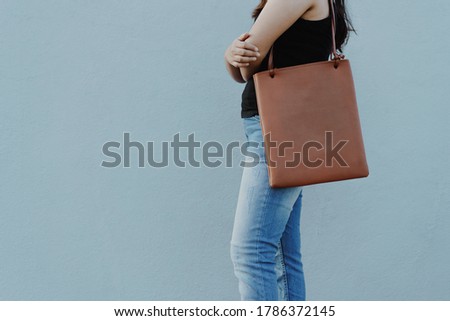 Brown leather tote bag on a young woman in blue jeans and tank top against grey background with copy space. Crop photo of woman folding hands standing casually