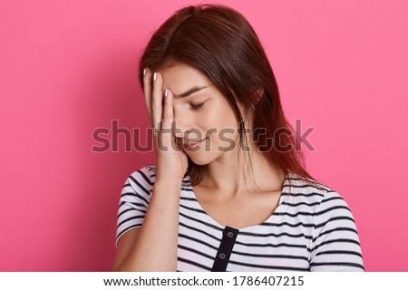 Tired young woman covers face with palm, closes her eyes, feels exhausted, wearing casual striped t shirt, posing isolated over pink background, wants to sleep.