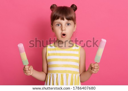 Girl with water ice creams in both hands wearing striped dress, having two knots, posing with astonished facial expression against pink background.