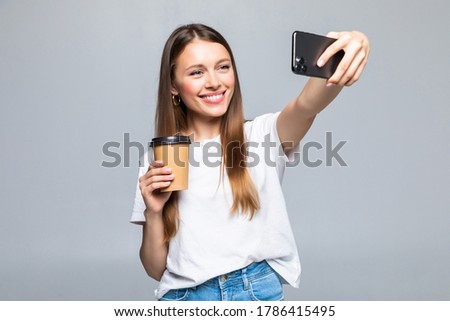 Portrait of woman taking selfie photo on smartphone in office and drinking takeaway coffee from plastic cup isolated over gray background