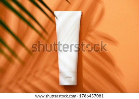 White plastic tube with face, hand and body cream on an orange background with palm leaves and shadow. Sun protection lotion, sunscreen. Summer skin care concept with spf flat lay. Mockup Royalty-Free Stock Photo #1786457081