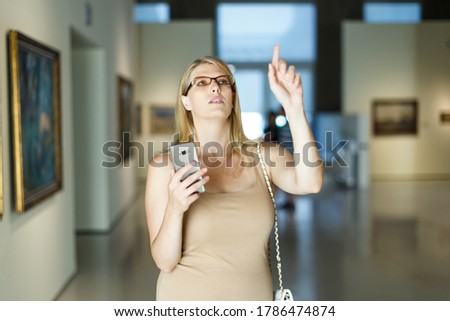 Positive woman wearing glasses with smartphone standing in hall of picture gallery and pointing to invisible screen