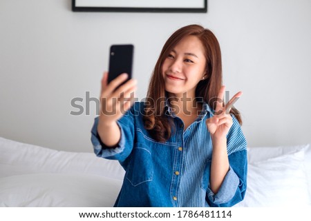Portrait image of a beautiful young asian woman using mobile phone to take selfie while sitting on a white bed