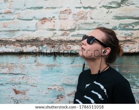Portrait of young attractive girl in black t-shirt and sunglasses in rock style on urban background listening to music with headphones. Hipster in wireless earphones on old blue brick wall backdrop.