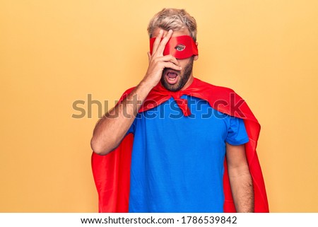 Handsome blond man wearing super hero costume with mask and cape over yellow background peeking in shock covering face and eyes with hand, looking through fingers afraid