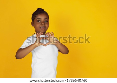 Inspired showing heart sign. Little african-american girl's portrait on yellow studio background. Cheerful kid. Concept of human emotions, facial expression, sales, ad. Copyspace. Looks cute.