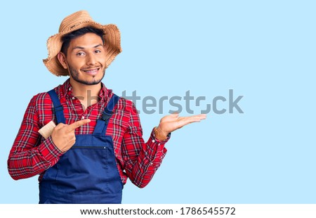 Handsome latin american young man weaing handyman uniform amazed and smiling to the camera while presenting with hand and pointing with finger. 