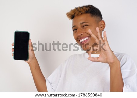 Excited African American woman showing smartohone blank screen, blinking eye and doing ok sign with hand. Studio shot of shocked girl holding smartphone with blank screen. Advertisement concept. Royalty-Free Stock Photo #1786550840