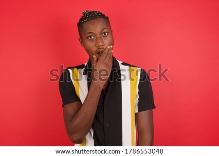 Emotional African american man with braids wearing casual shirt over isolated red background gasps from astonishment, covers opened mouth with palm, looks shocked at camera. Reaction concept.