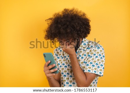 A beautiful Young man with afro hair over  wearing hawaiian shirt standing over yellow background looking at smart phone feeling sad holding hand on face.
