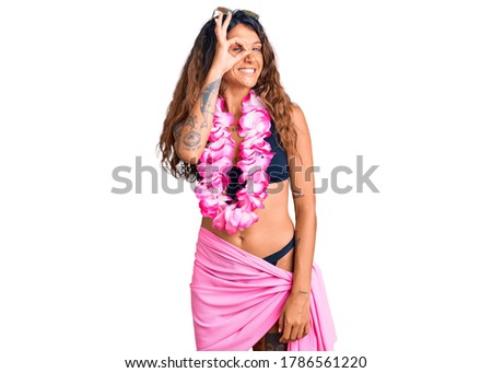 Young hispanic woman with tattoo wearing bikini and hawaiian lei smiling happy doing ok sign with hand on eye looking through fingers 