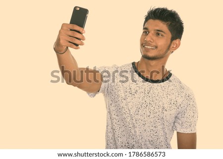 Studio shot of young happy Indian man smiling while taking selfie picture with mobile phone