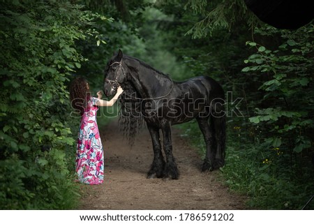 Young brunette woman in a bright dress posing with a frieze horse in a green forest.