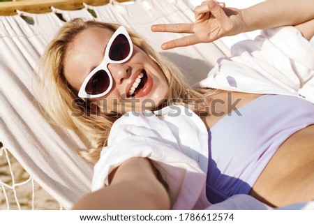 Image of smiling woman gesturing peace gesture and taking selfie photo while lying in hammock on summer beach
