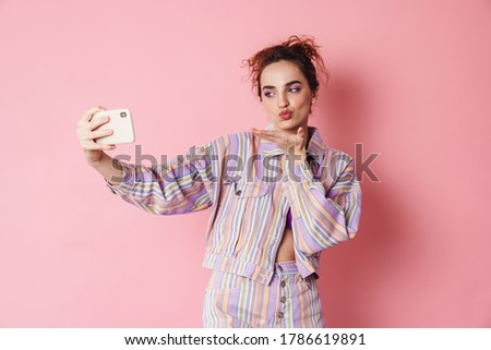 Image of ginger happy woman blowing air kiss while taking selfie on cellphone isolated over pink background