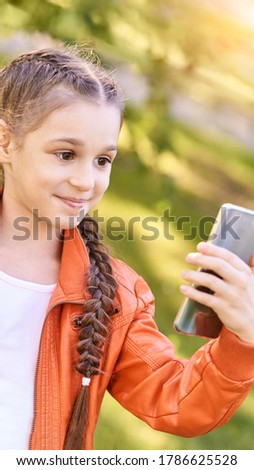 Little girl doing selfie. Happy pretty kid. Green outdoor background. Orange dress. Female person self photo. Schoolgirl phone. Travel activity. Stacation teenage influencer