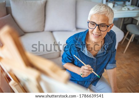 Portrait of attractive middle aged professional female painter with gray hair working at home standing in front of easel, having thoughtful look, finishing still life painting Royalty-Free Stock Photo #1786639541