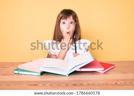 Little caucasian kid girl with long hair studying for school exam covering mouth with hand, shocked and afraid for mistake. surprised expression 