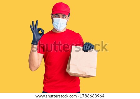 Young hispanic man delivering food wearing covid-19 safety mask holding paper bag doing ok sign with fingers, smiling friendly gesturing excellent symbol 