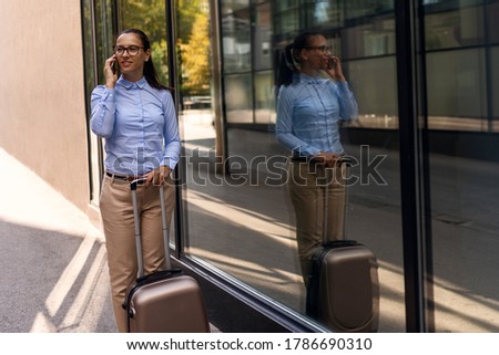 Handsome young woman on business trip walking with her luggage and talking on cellphone at airport. Travelling business woman making phone call.


