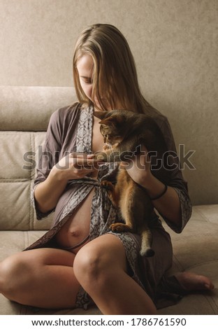 pregnant girl is sitting at home on the beige sofa with cat in hands and smiling cute. lifestyle concept, free space