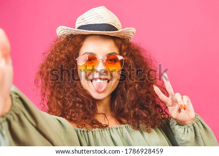 Young beautifult girl or woman making selfie in studio and laughing. Good-looking young woman with curly hair taking picture of herself over isolated pink background.