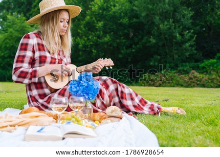 Girl in red checkered dress and hat sitting on white knit picnic blanket plays ukulele and drinking wine. Summer picnic on sunny day with bread, fruit, bouquet hydrangea flowers. Selective focus