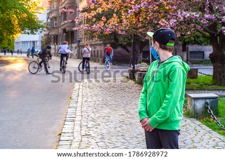 A lonely boy in a protective mask watches a group of teenagers on bicycles. Quarantine coronavirus covid-19. Outcast in the company. Sad lonely teen looks at teenagers with envy