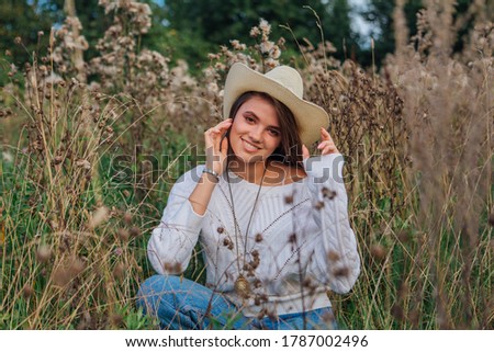 Young beautiful brunette woman dressed in a white sweater, jeans and cowboy straw hat sitting in dry brown bur grass, smiling and laughing during sunset