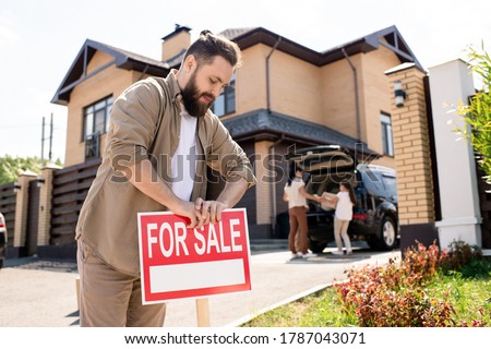 Young bearded father placing For sale sign into ground near house while putting house up for sale