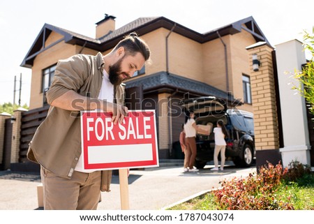 Serious bearded man in casual shirt placing For sale sign against house to pay attention of buyers