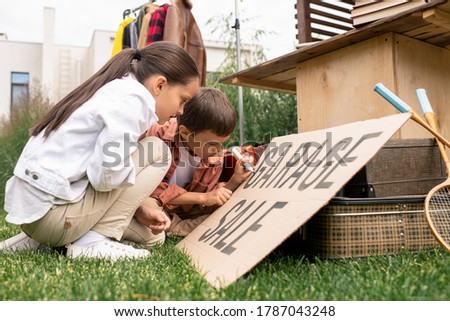 Creative cute kids sitting on grass and making garage sale sign together in backyard