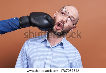 Funny grimmasa, a young bald businessman in a blue shirt and glasses, gets an unexpected blow in the face with a hand in a black boxer glove on the right side. The glasses had slipped from his face.