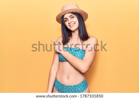 Young beautiful woman wearing bikini and summer hat cheerful with a smile of face pointing with hand and finger up to the side with happy and natural expression on face 