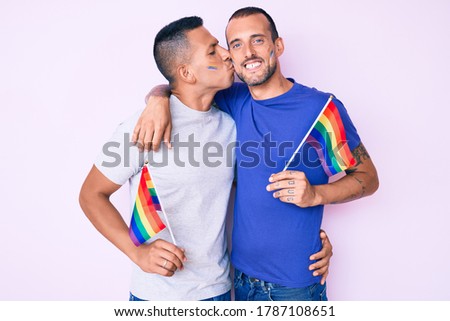 Young couple of gay men together in love holding homosexual pride colorful flag kissing with affection