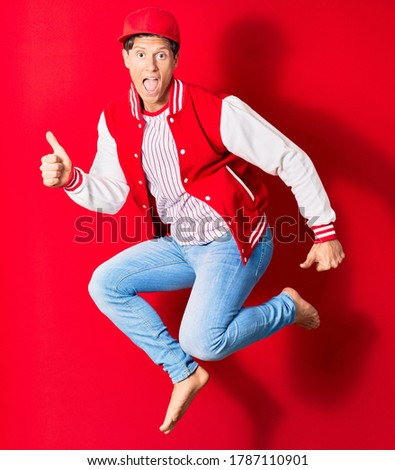 Young handsome man wearing baseball clothes doing ok sing with thums up. Jumping with open mouth over isolated red background